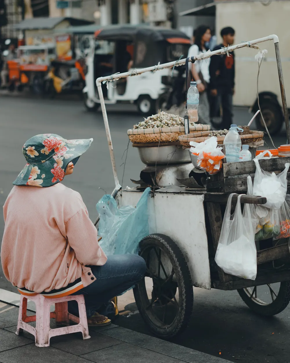 A street food vendor beside their cart on a Phnom Penh street, with tuk-tuks passing behind.